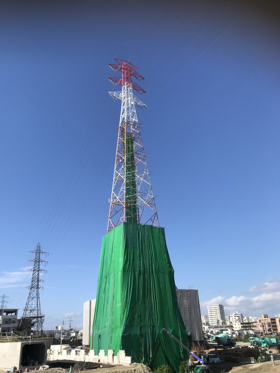 青空の下、緑の養生メッシュに覆われた紅白鉄塔の全景